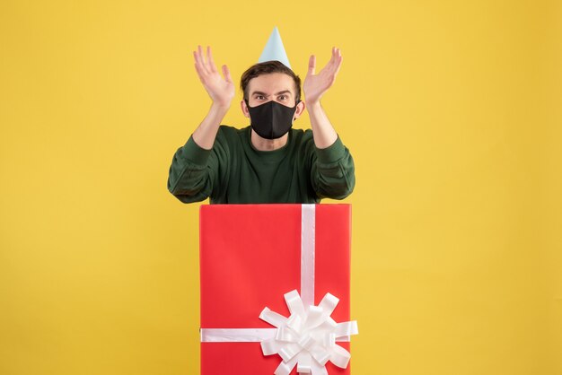 Front view angry young man with party cap and mask standing behind big giftbox on yellow 