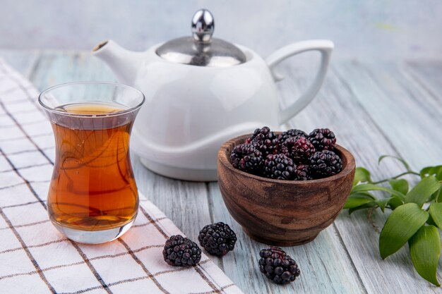 Front view of armudu glass of tea with teapot and blackberry on a gray surface