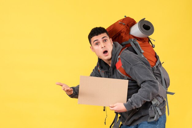 Вид спереди изумленный молодой человек с красным рюкзаком, держащим картон