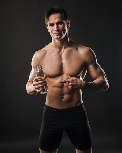 Front view of athletic shirtless man pointing at water bottle