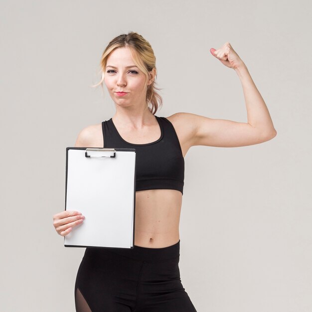 Front view of athletic woman posing while showing bicep and holding notepad