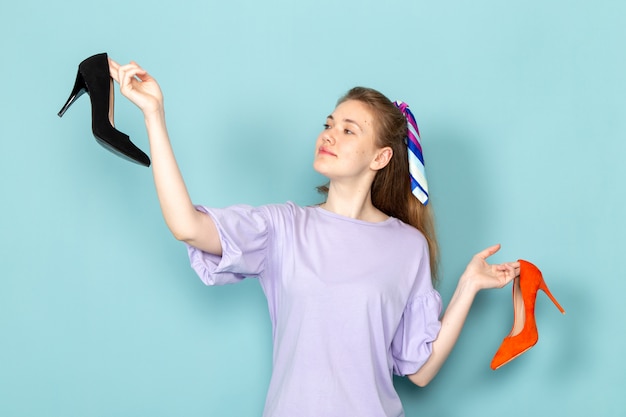 A front view attractive female in blue shirt-dress holding black and orange shoes on blue