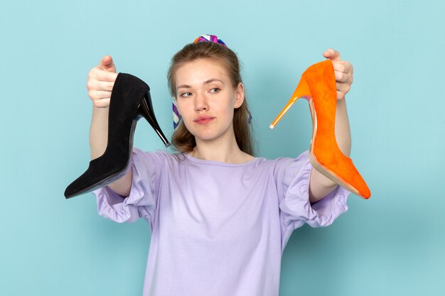 A front view attractive female in blue shirt-dress holding black and orange shoes on blue