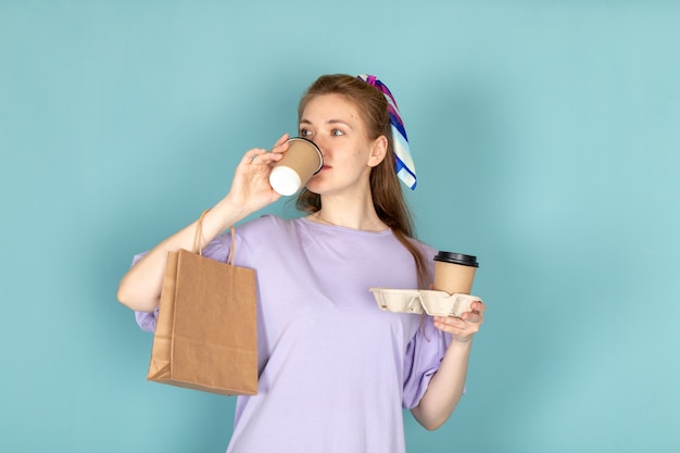 A front view attractive female in blue shirt-dress holding paper package and coffee cups drinking on blue