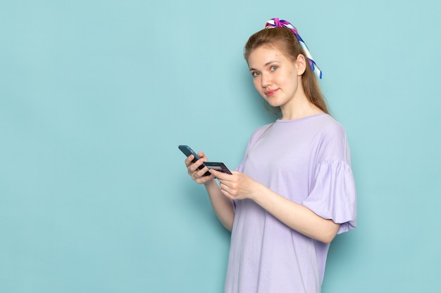 A front view attractive female in blue shirt-dress holding and using a phone smiling on blue