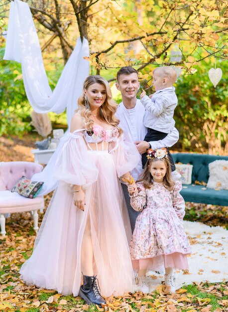 Front view of attractive woman with long curly hair dressed in pink dress with long sleeves holding her little daughter by hands