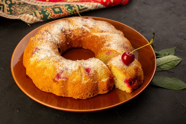 A front view baked fruit cake delicious round with red cherries inside and sugar powder inside round brown plate on dark