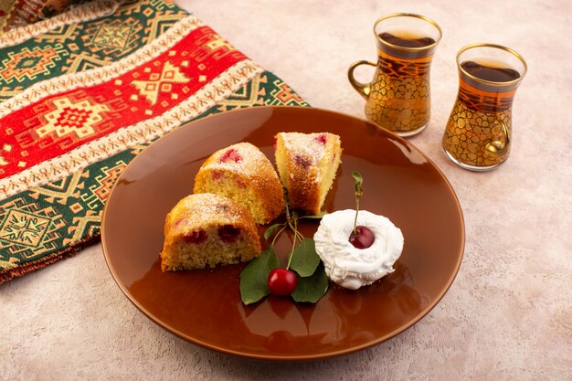 A front view baked fruit cake delicious sliced with red cherries inside and sugar powder inside round brown plate on pink