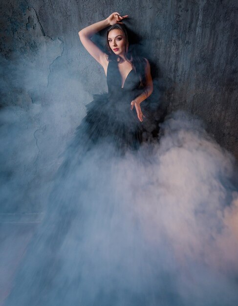 Front view of beautiful brunette in a black dress all in smoke posing on the background of the wall