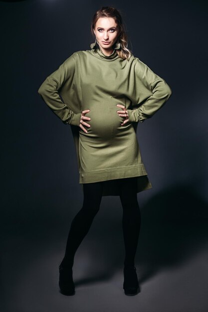 Front view of beautiful and fashionable pregnant brunette woman at dark studio Pretty and stylish woman wearing in khaki dress holding pregnant belly looking at camera Concept of pregnancy