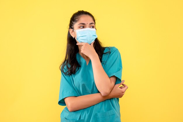 Front view beautiful female doctor in uniform putting hand to her chin standing on yellow background