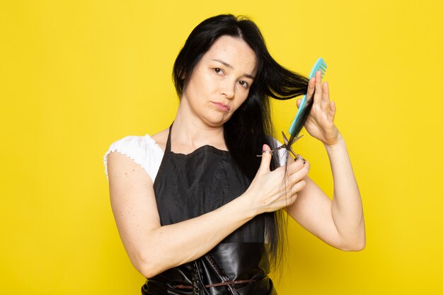 A front view beautiful female hairdresser in white t-shirt black cape with brushes with washed hair brushing and cutting her hair posing on the yellow background stylist barber hair