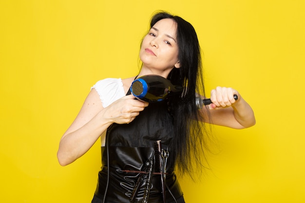 A front view beautiful female hairdresser in white t-shirt black cape with brushes with washed hair drying brushing her hair posing on the yellow background stylist barber hair