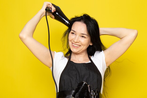 A front view beautiful female hairdresser in white t-shirt black cape with brushes with washed hair drying her hair posing smiling on the yellow background stylist barber hair