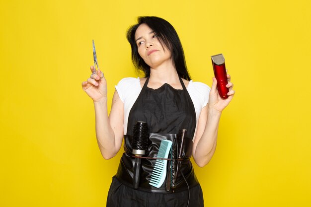 A front view beautiful female hairdresser in white t-shirt black cape with brushes with washed hair holding scissors and machine posing on the yellow background stylist barber hair