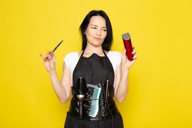 A front view beautiful female hairdresser in white t-shirt black cape with brushes with washed hair holding scissors and machine posing on the yellow background stylist barber hair