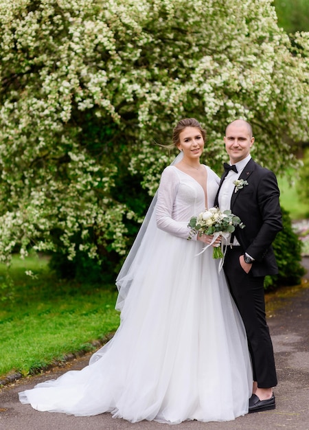 Front view of beautiful girl with stylish hairdo and make up wearing in long wedding dress and veil holding flowers posing with handsome groom among amazing plants and together looking at camera