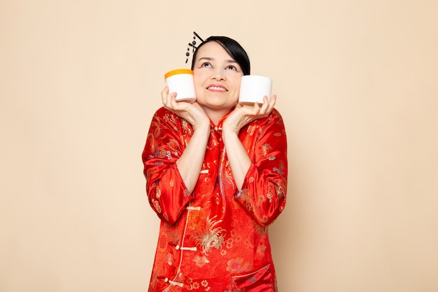 Free photo a front view beautiful japanese geisha in traditional red japanese dress with hair sticks posing holding cream cans smiling on the cream background ceremony japan