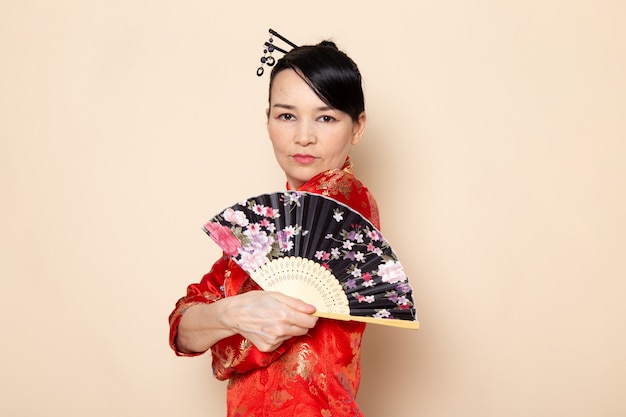 A front view beautiful japanese geisha in traditional red japanese dress with hair sticks posing holding folding fan elegant on the cream background ceremony japan