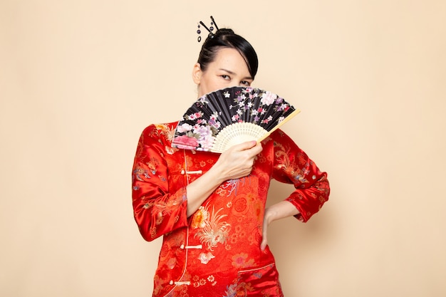 A front view beautiful japanese geisha in traditional red japanese dress with hair sticks posing holding folding fan elegant on the cream background ceremony japan