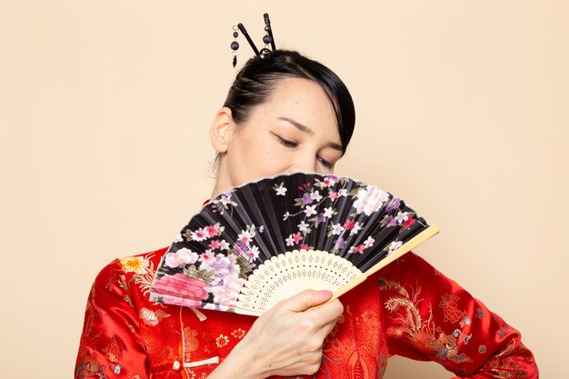 A front view beautiful japanese geisha in traditional red japanese dress with hair sticks posing holding folding fan smiling on the cream background ceremony japan