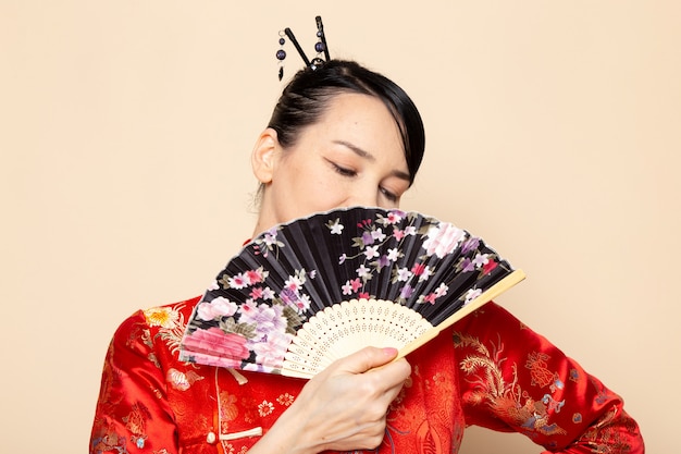 正面を描いた伝統的な赤い和服で美しい日本の芸者のヘアスティックでポーズを保持している扇子をクリーム色の背景に笑みを浮かべて式日本