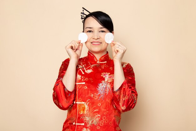 A front view beautiful japanese geisha in traditional red japanese dress with hair sticks posing holding little white cotton elegant smiling on the cream background ceremony japan