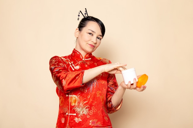 A front view beautiful japanese geisha in traditional red japanese dress with hair sticks posing opening cream can smelling on the cream background ceremony japan