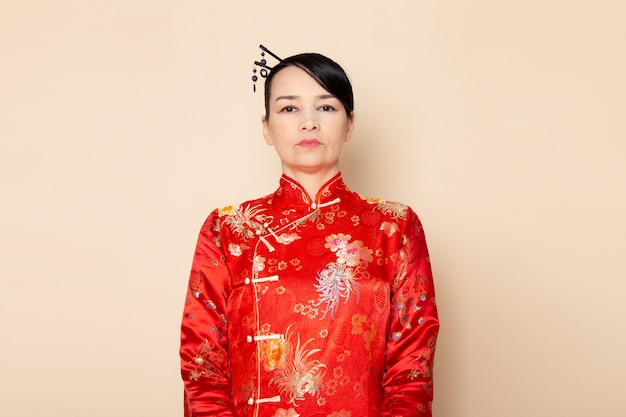 正面を飾るクリーム色の背景の上に立ってポーズをとって髪棒で伝統的な赤い和服の美しい日本の芸者