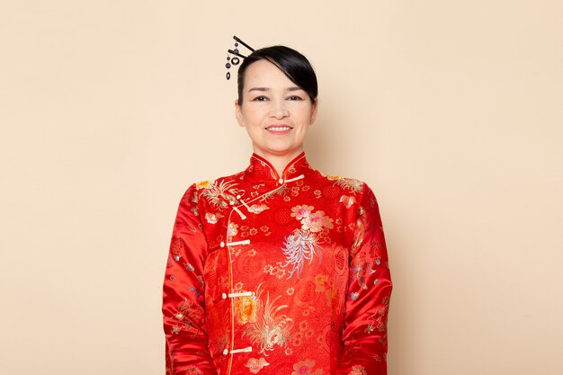 Вид спереди красивая японская гейша в традиционном красном японском платье с заколками для волос, стоя на кремовом фоне, улыбающаяся церемония, занимательная япония восток