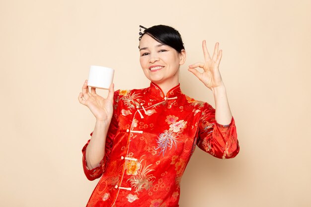 クリームを使用してポーズをとって髪棒でポーズをとって伝統的な赤い和服で正面の美しい日本の芸者は、クリーム色の背景に笑うことができます。