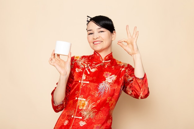 Вид спереди красивая японская гейша в традиционном красном японском платье с заколками для волос позирует в кремовой банке улыбается на кремовом фоне церемонии японии