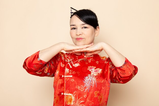 正面東日本を楽しませるクリーム色の背景の上に立って彼女の手でポーズをとって髪棒で伝統的な赤い和服で美しい日本の芸者