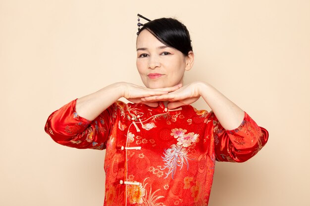 A front view beautiful japanese geisha in traditional red japanese dress with hair sticks posing with her hands standing on the cream background ceremony entertaining japan east