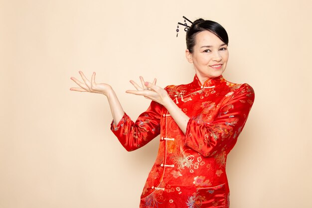Вид спереди красивая японская гейша в традиционном красном японском платье с заколками для волос позирует руками, стоя, улыбаясь на кремовом фоне, церемония занимательная япония восток