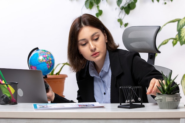 A front view beautiful young businesswoman in black jacket and blue shirt observing little globe in front of table business job office