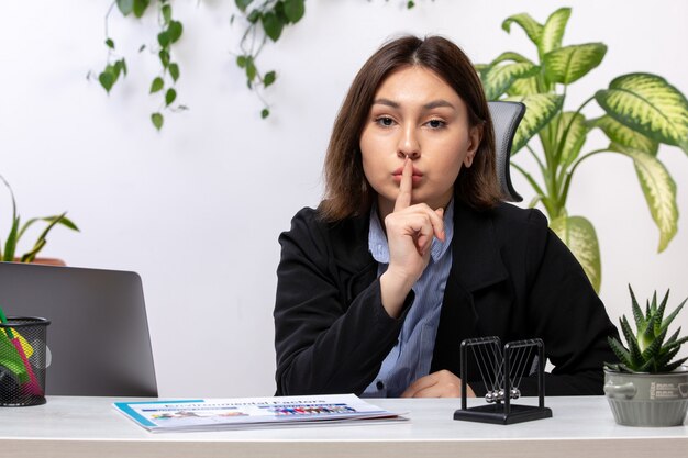 A front view beautiful young businesswoman in black jacket and blue shirt showing silence sign in front of table business job office