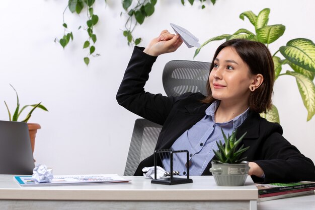 A front view beautiful young businesswoman in black jacket and blue shirt smiling throwing paper planes in front of table business job office