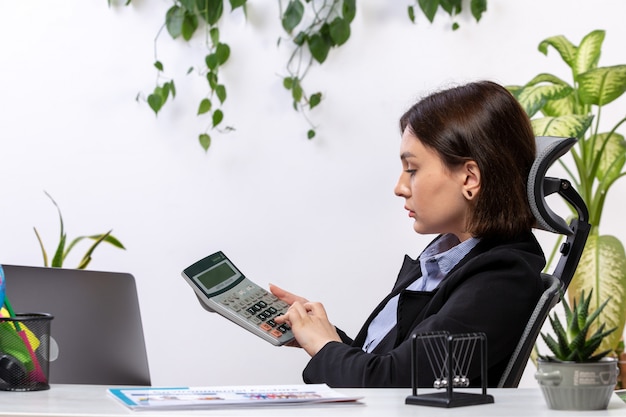 A front view beautiful young businesswoman in black jacket and blue shirt working with calculator in front of table business job office