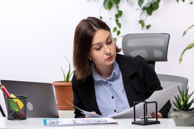 A front view beautiful young businesswoman in black jacket and blue shirt working with laptop and documents in front of table business job office