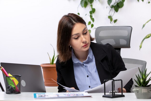 A front view beautiful young businesswoman in black jacket and blue shirt working with laptop and documents in front of table business job office