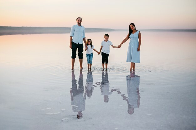 Вид спереди красивых молодых родителей, их милая маленькая дочь и сын, держась за руки, гуляют по пляжу