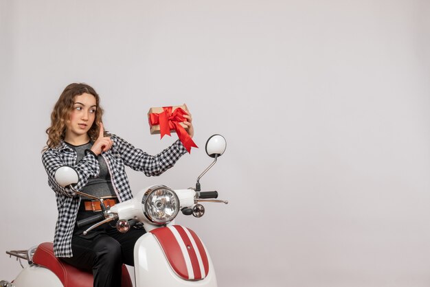 Front view of beautiful young woman on moped holding gift on grey wall