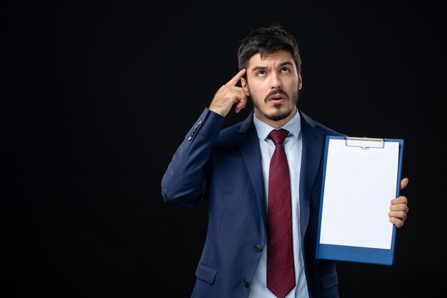 Front view of bewildered adult in suit holding free space for writing and pointing something on the right side on isolated dark wall