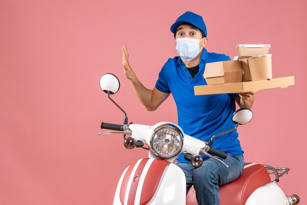 Front view of bewildered male delivery person in mask wearing hat sitting on scooter delivering orders on pastel peach background