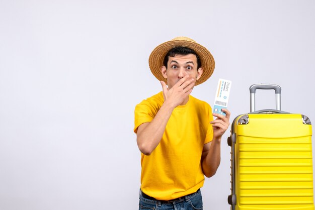 Front view bewildered young tourist standing near yellow suitcase holding ticket