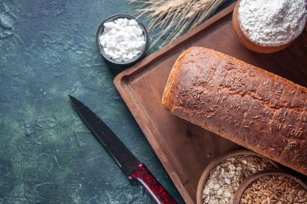 Front view of black bread slices flour in a bowl on wooden board and knive spikes raw oatmeal wheat on the left side on mixed colors distressed background