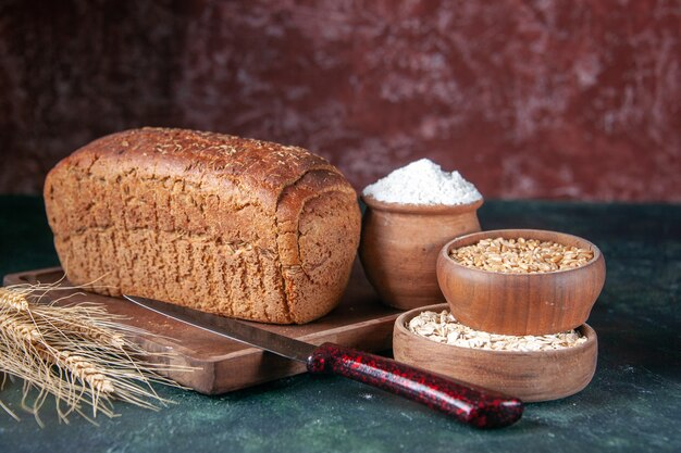 Front view of black bread slices flour in a bowl on wooden board and knive spikes raw oatmeal wheat on mixed colors distressed background