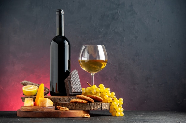 Вид спереди бутылка черного вина красное вино в стеклянном сырном разрезе лимон кусок темного шоколадного печенья виноград на деревянных досках на темно-красном столе с местом для копирования