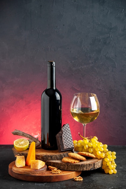 正面図ガラスチーズカットレモンの黒ワインボトル赤ワインダークレッドテーブルの上の木の板にダークチョコレートビスケットブドウの一部